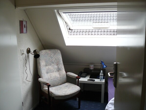 A small room with a slanted ceiling, a single chair, a table with a phone, a remote control, and a water bottle.