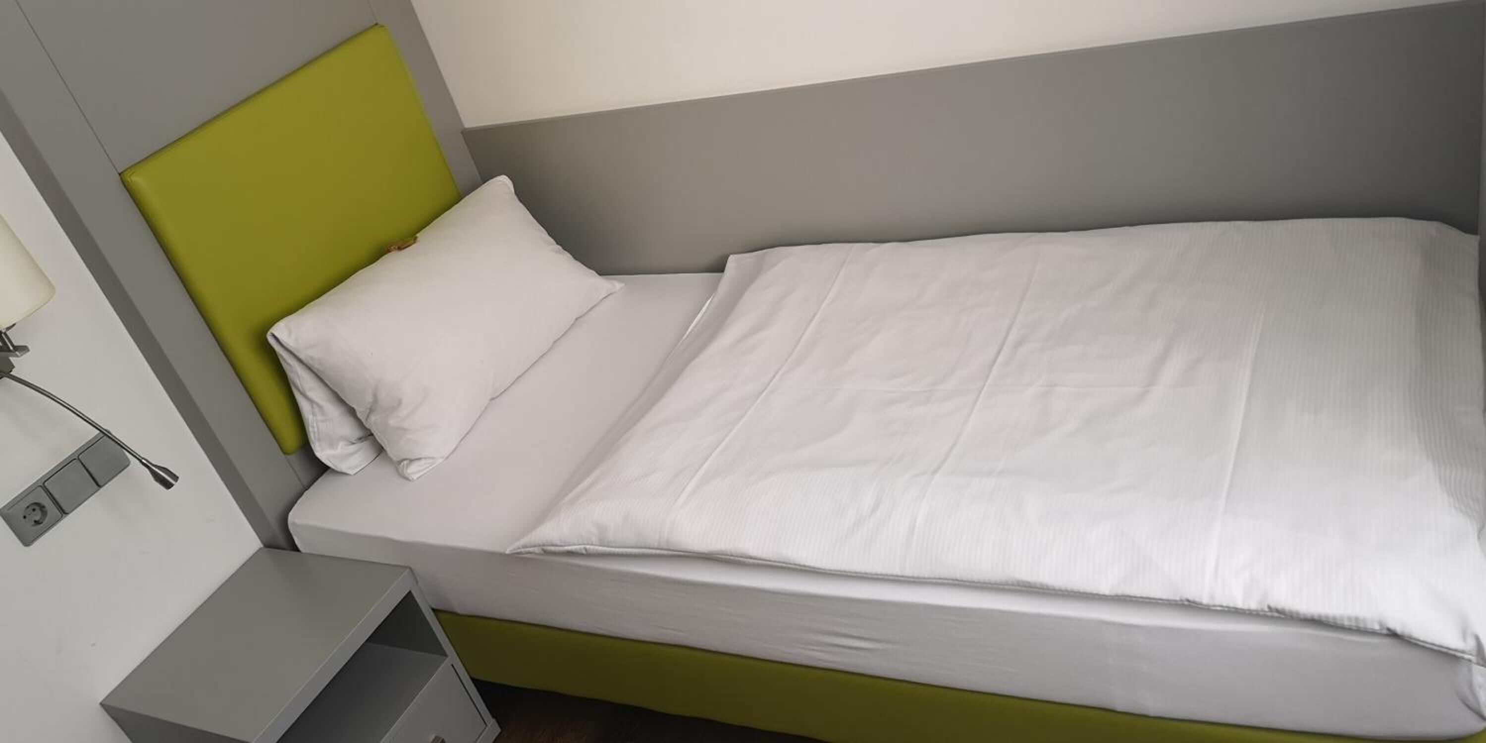 A single-bed room with a green headboard, white bedding, and a small bedside table with a lamp.