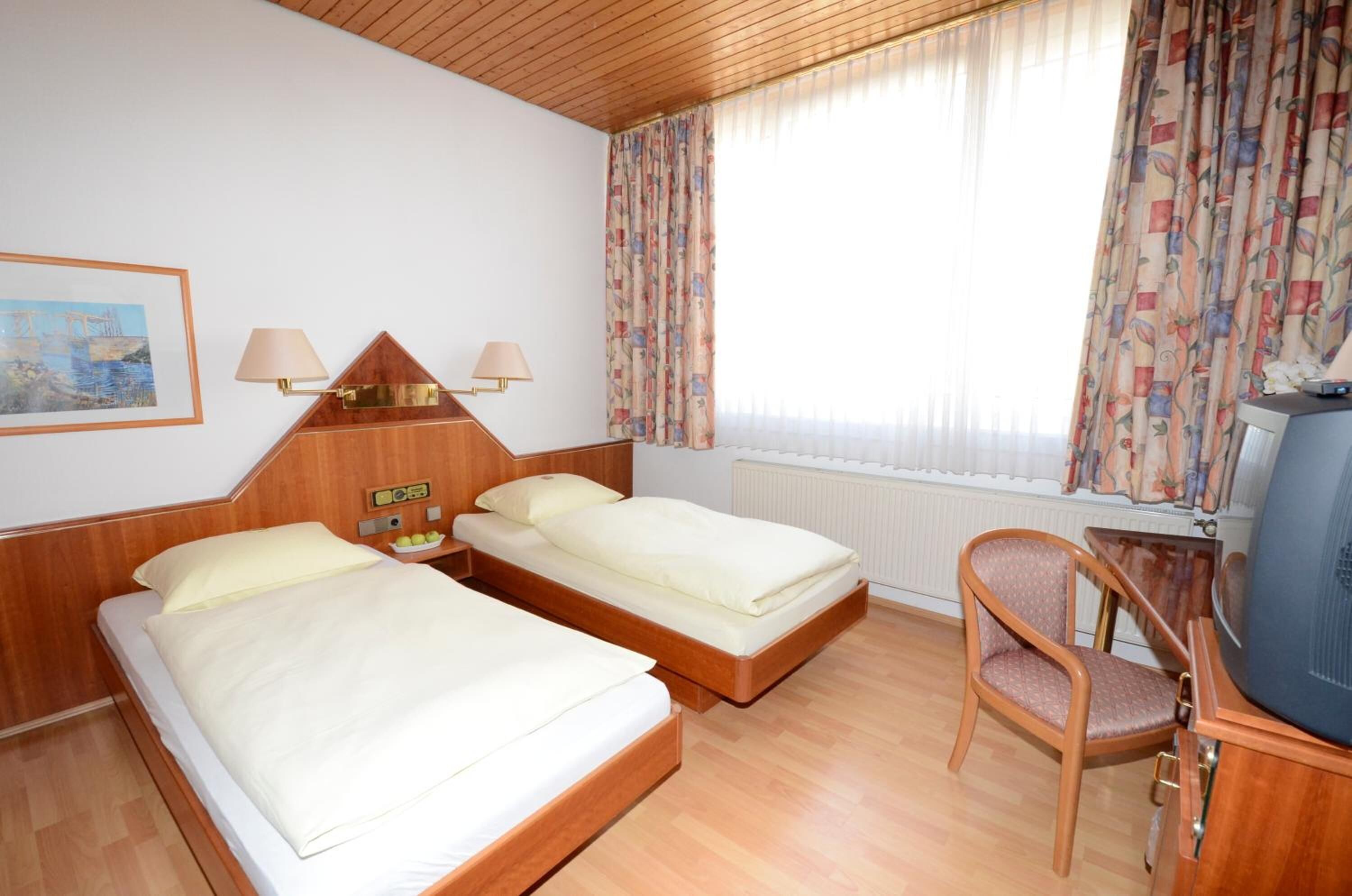 Ein Hotelzimmer mit zwei Betten, einem Schreibtisch, einem Fernseher, einem Sessel und einem Fenster mit Vorhängen.