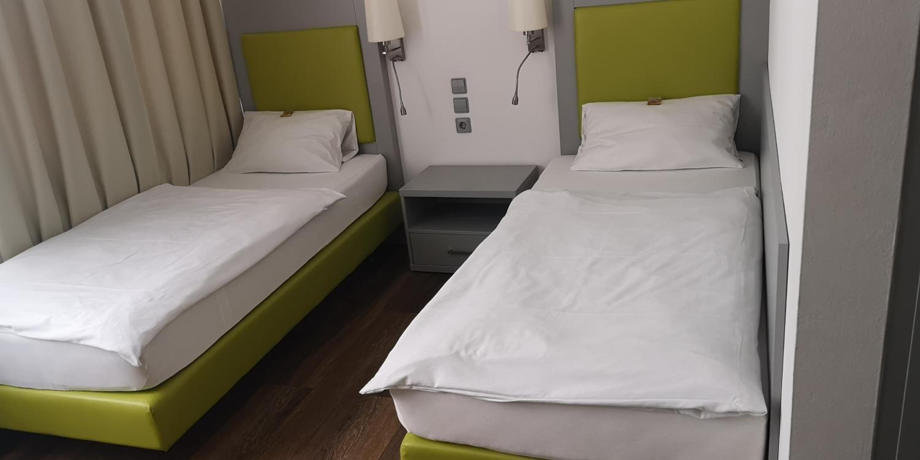 Ein Hotelzimmer mit zwei Einzelbetten, jeweils mit einem grünen Kopfteil, weißer Bettwäsche und einem Nachttisch mit einer Lampe.