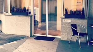 A building entrance with a glass door, potted plants, and a chair outside.