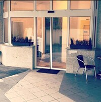 Entrada de um edifício com porta de vidro, plantas em vasos e uma cadeira do lado de fora.
