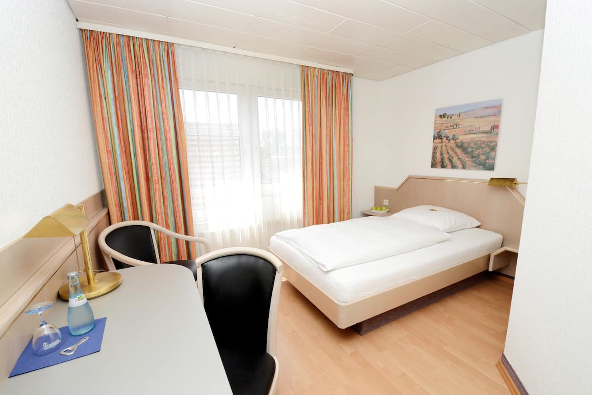 A hotel room with a bed, a desk with a lamp, a chair, a window with curtains, and a painting on the wall.
