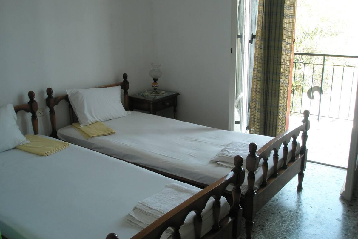 A double bed with wooden headboard and footboard, a bedside table with a lamp, and a window with a view of greenery outside.