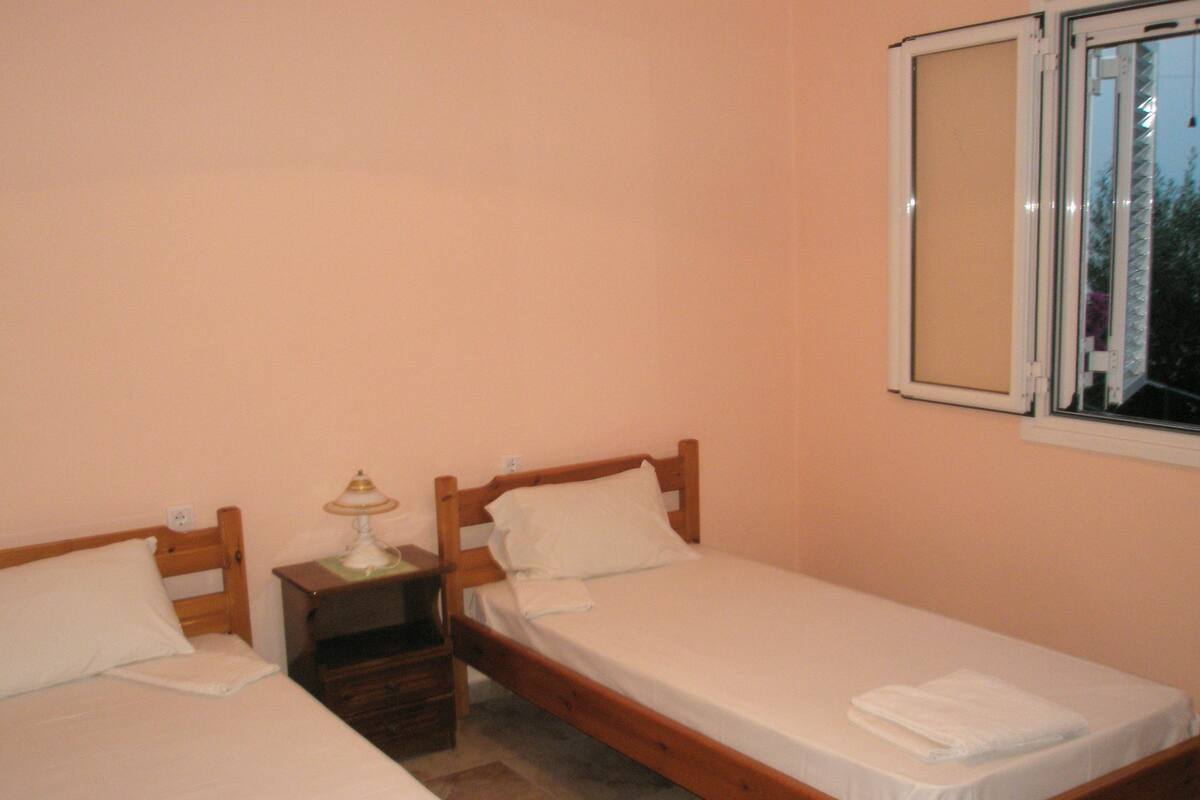 A bedroom with two single beds, a nightstand with a lamp, and a window.