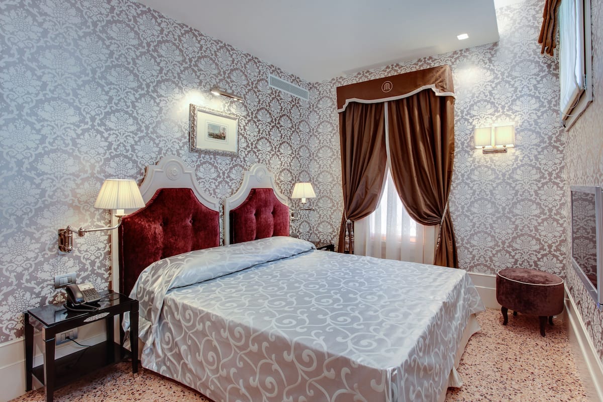 Une chambre avec un grand lit, deux lampes de chevet, une table de chevet avec un téléphone, un petit pouf rond et une fenêtre avec des rideaux.