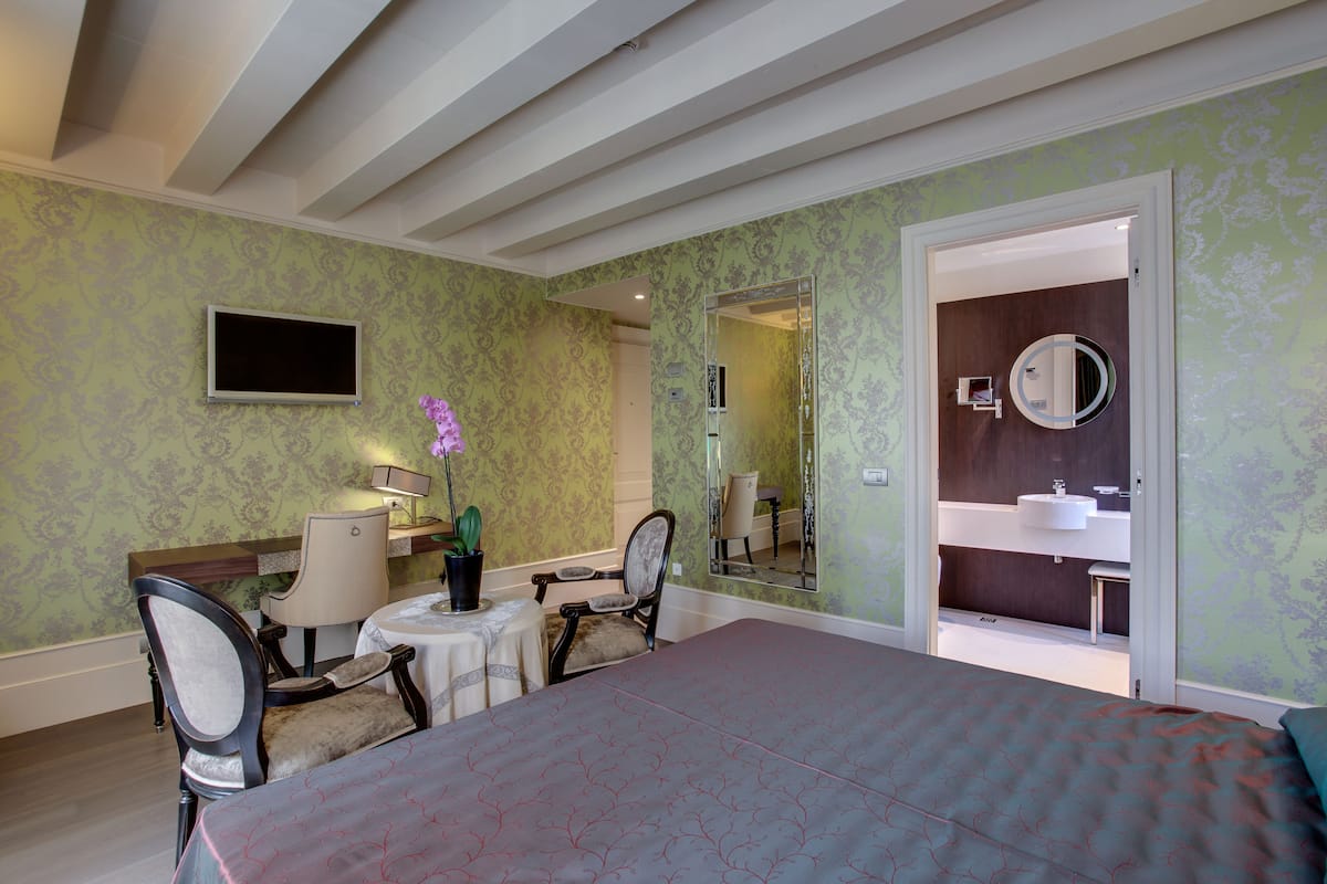 Une chambre d’hôtel avec un lit, deux chaises, une petite table, une plante en pot, un téléviseur et une salle de bain visible à travers une porte ouverte.