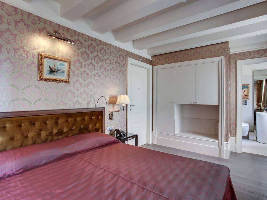 Une chambre d’hôtel avec un grand lit, une tête de lit, une table de chevet avec une lampe, un tableau encadré au mur et une armoire.