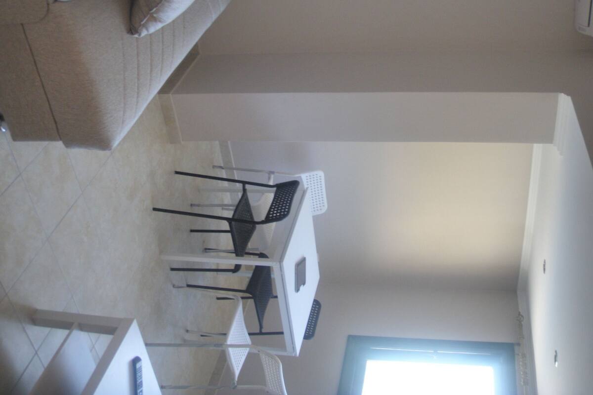 Un área de comedor con una mesa y sillas blancas, un sofá y una ventana con luz natural.