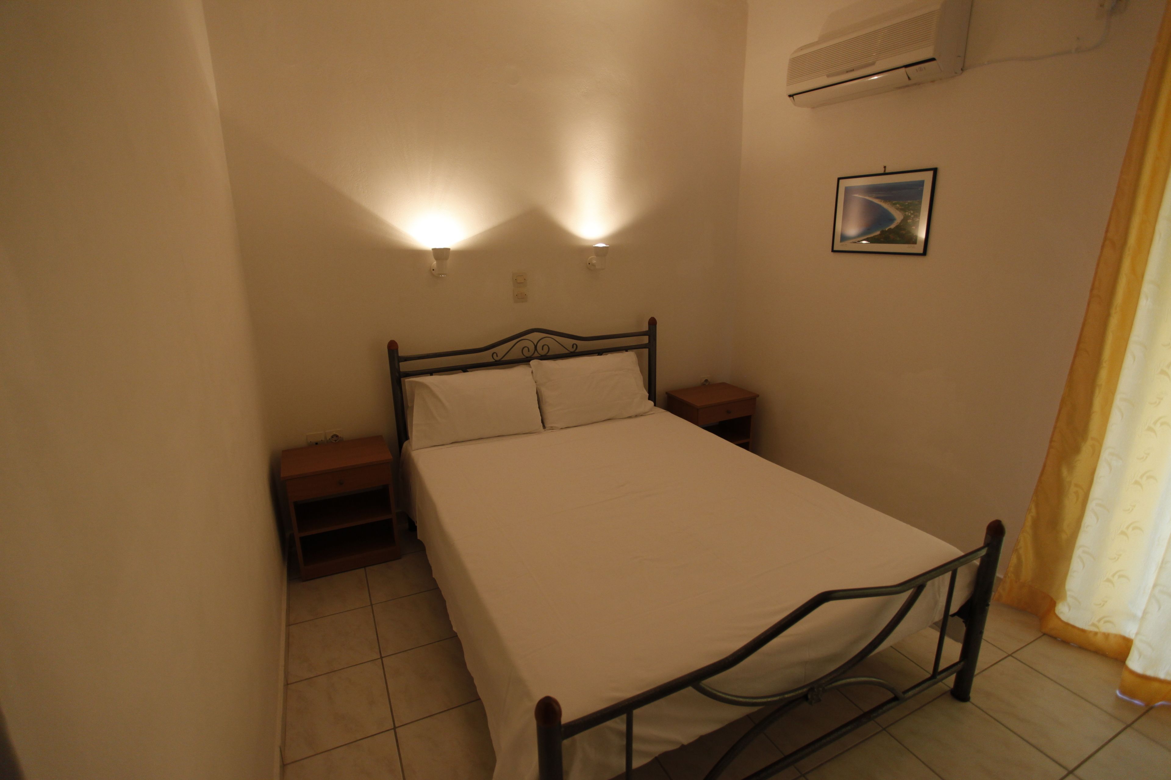 A bedroom with a bed, two bedside lamps, a nightstand, a wall-mounted air conditioner, and a framed picture.