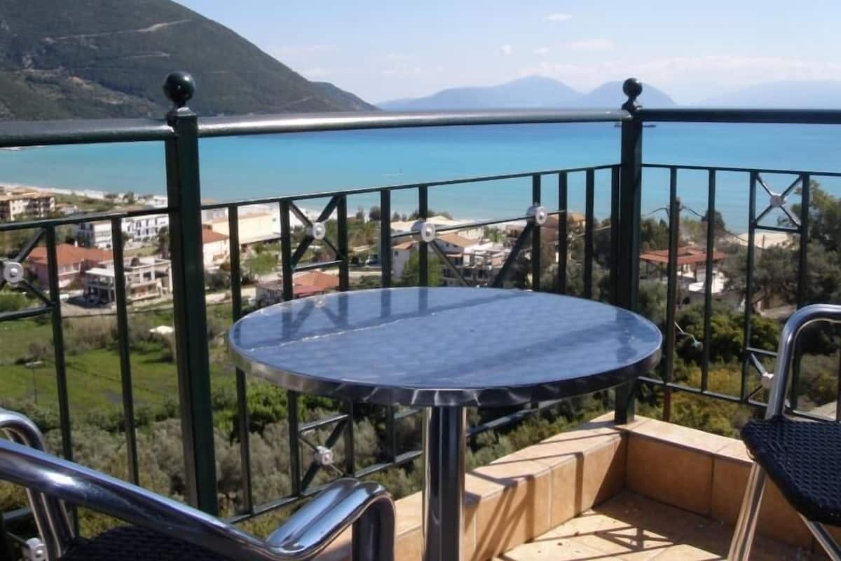 Un balcón con una mesa redonda y sillas, con vistas a una zona costera con edificios y montañas.