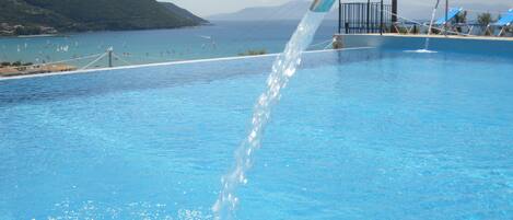 A swimming pool with clear blue water, a water fountain, and a view of the sea and hills in the background.