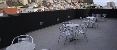 Una terraza en la azotea con mesas y sillas de metal, con vistas a un pueblo en la ladera densamente poblado.