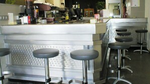 Uma área de bar moderna com balcões e banquetas de aço inoxidável, equipada com diversos utensílios de bar e garrafas.