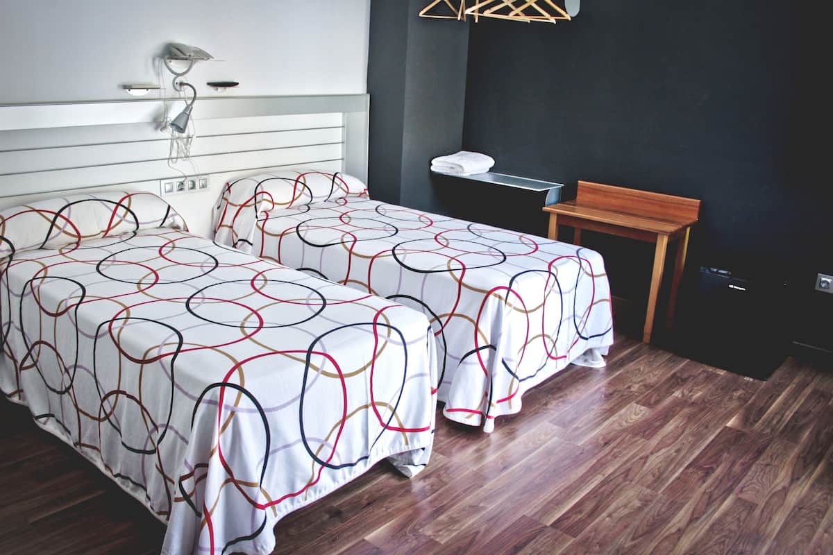 A hotel room with two beds, a wooden bench, a desk, and a wall-mounted lamp.