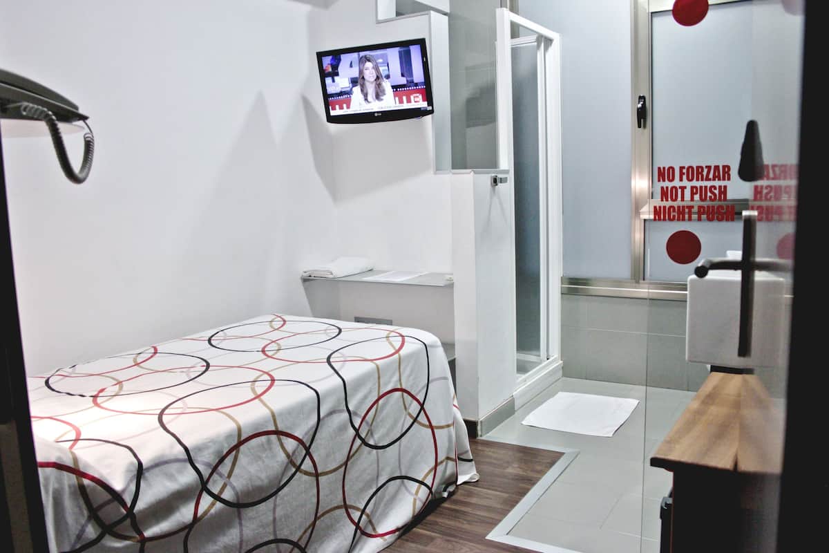 A hotel room with a bed, a TV mounted on the wall, a shower area, and a sign with instructions.