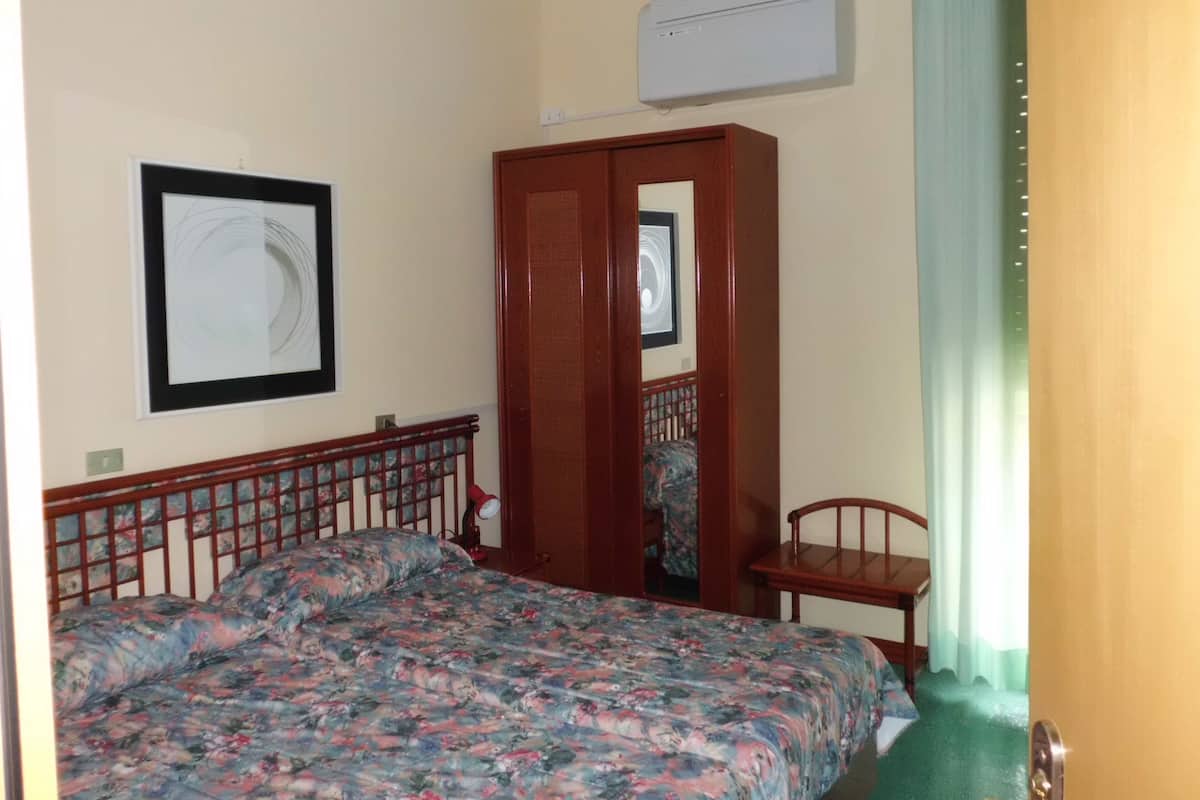 Un dormitorio con cama, armario, silla, un cuadro en la pared y equipo de aire acondicionado.