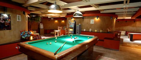 Una mesa de billar con superficie de fieltro verde, rodeada por una decoración en tonos rojo y blanco, y una zona de bar con taburetes y una caja registradora.