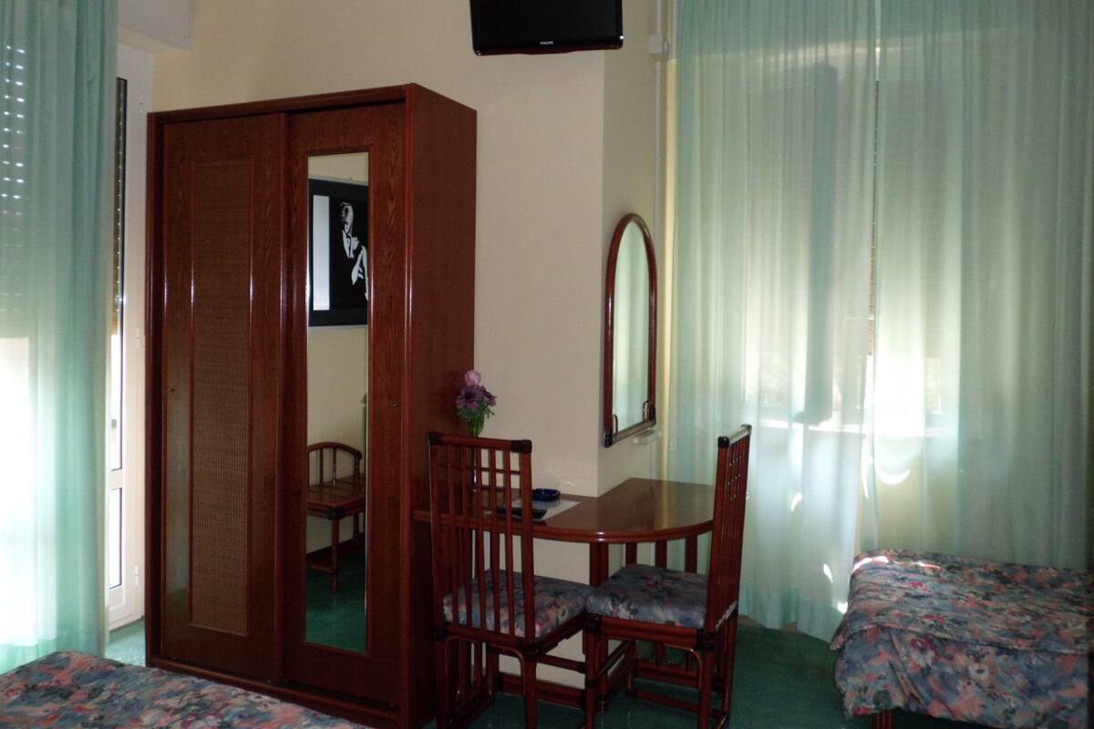 Habitación de hotel con un armario de madera, una mesita con una silla, una cama, un espejo y un televisor en la pared.
