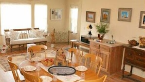 Un comedor con una mesa redonda preparada para cuatro personas, rodeada de sillas de madera. Se observa un sofá y una televisión al fondo.