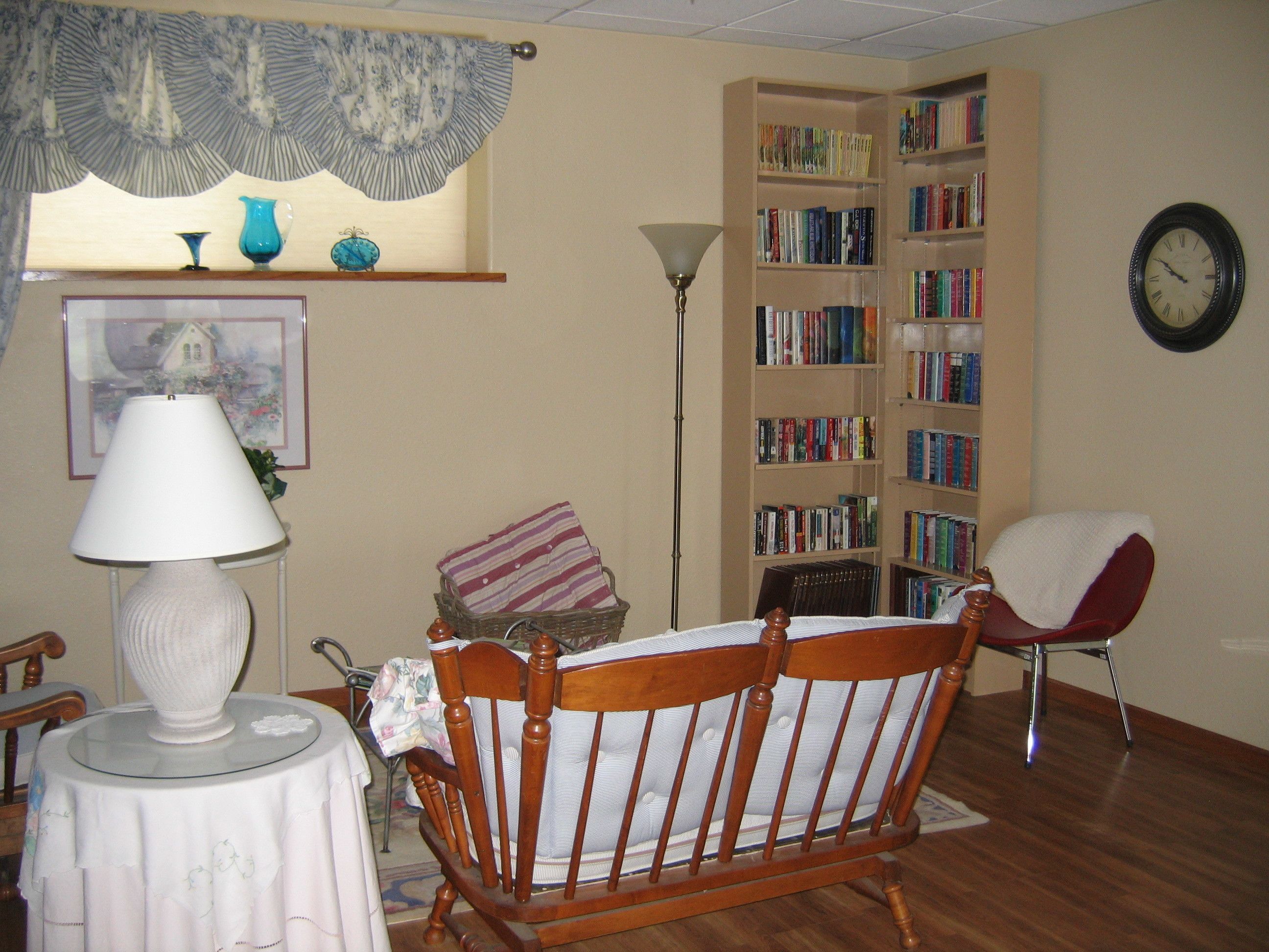 Un salon avec un fauteuil en bois, une table avec une lampe, une bibliothèque et une horloge au mur.