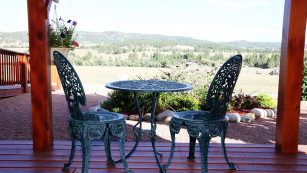 Una terraza de madera con dos sillas de metal decorativas y una mesa redonda, con vistas a un paisaje pintoresco.