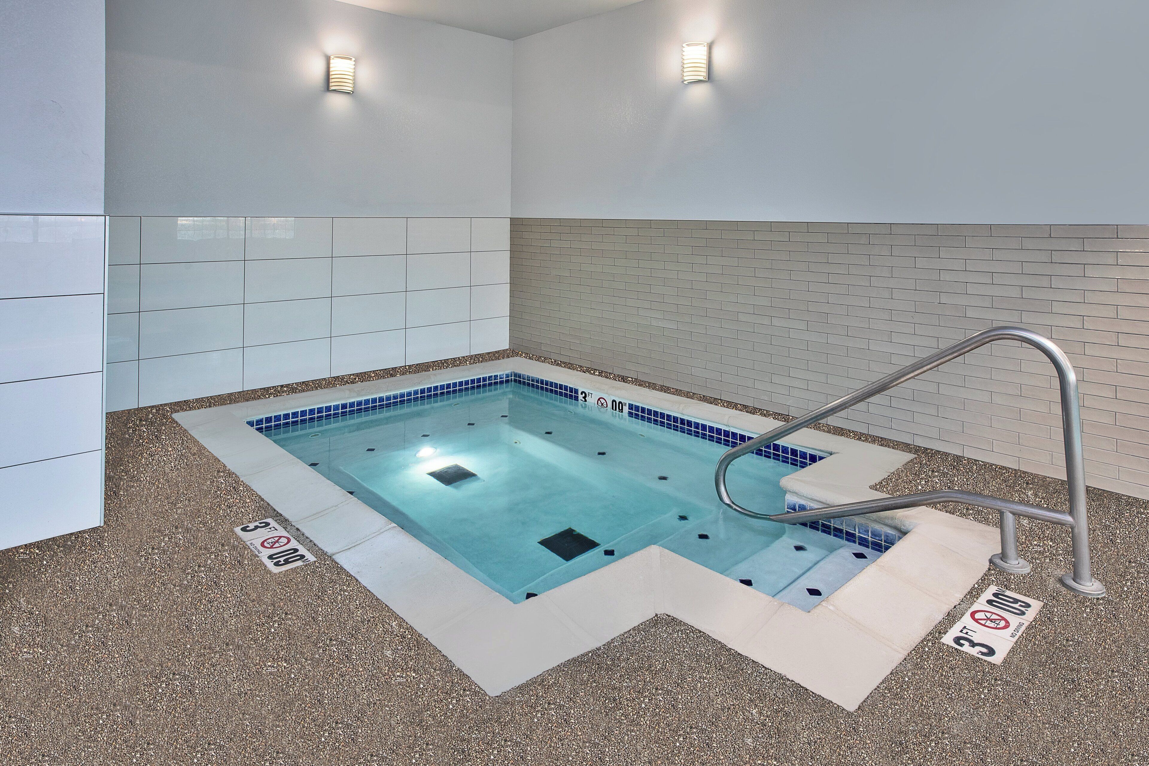 Una piscina interior pequeña y rectangular con base de azulejos azules y pared blanca que la rodea. Hay dos barandillas de metal en el borde de la piscina, y un letrero en el suelo que indica una profundidad de 3 pies.