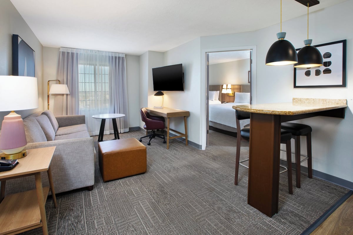 A hotel room with a living area, a desk, a bed, a TV, and a kitchenette.