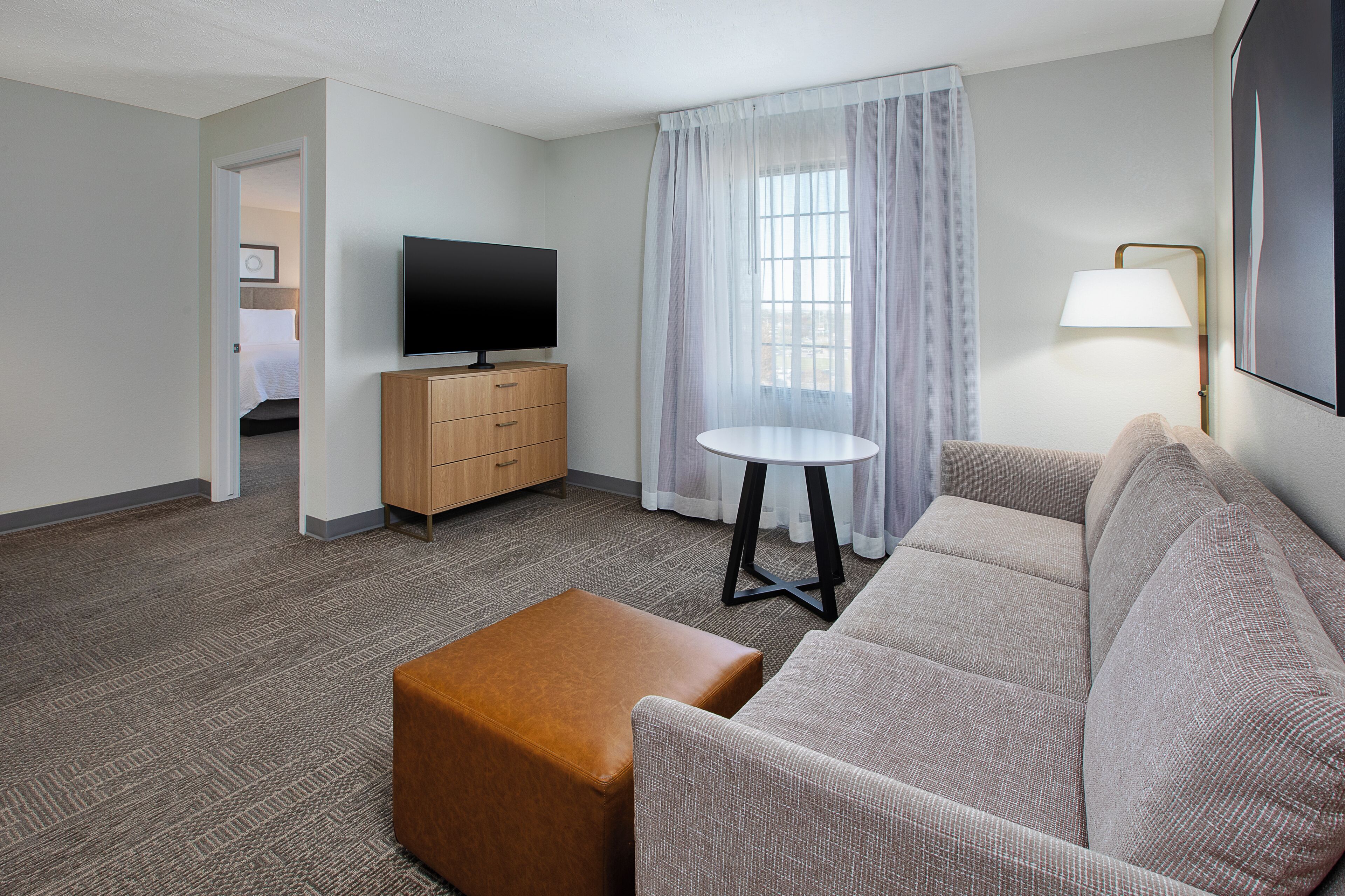 A hotel room with a sofa, ottoman, flat-screen TV, wooden cabinet, and a window with sheer curtains.