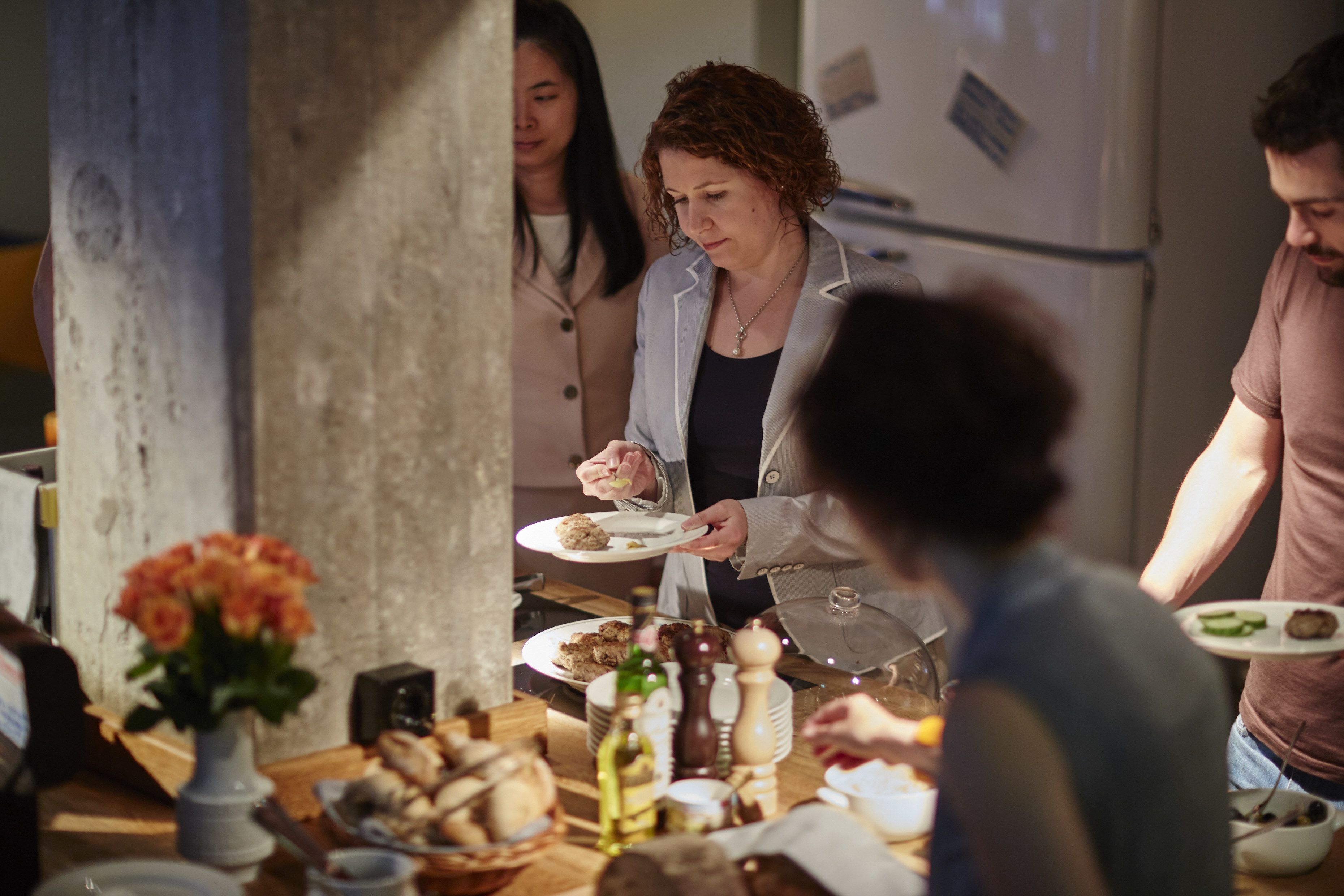 A group of people around a table with food and drinks, in a room with a concrete pillar and a refrigerator in the background.