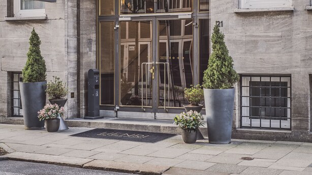 Het uiterlijk van Hotel Henri, met een grote glazen entree, een naambord en potplanten.