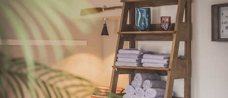 Una estantería de madera con toallas dobladas y un florero, una silla estampada y un revistero.