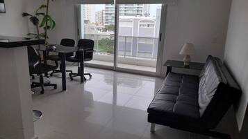 A modern living room with a black sofa, a dining table with chairs, a small table with a lamp, and a large window with a view of buildings.