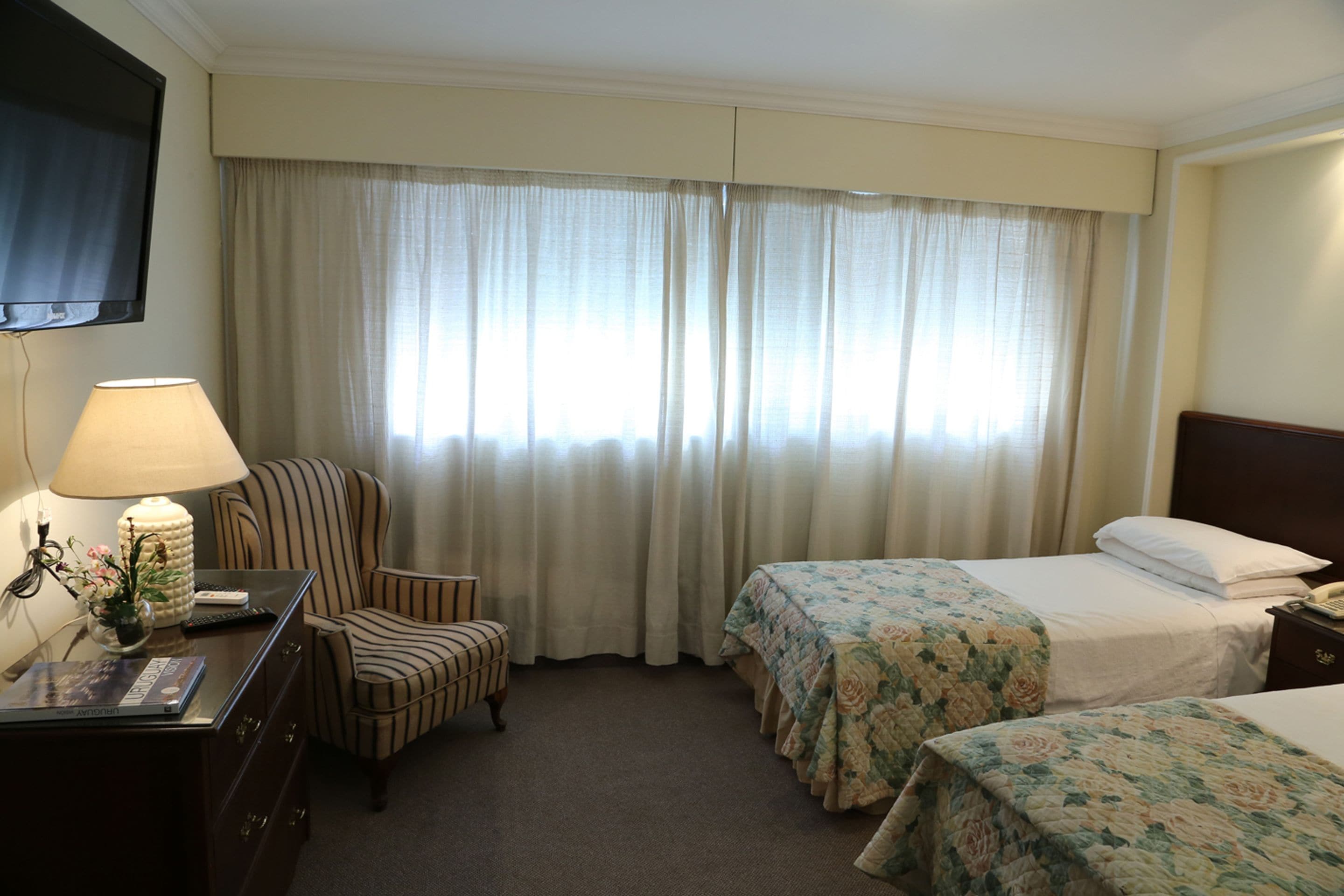 A hotel room with two beds, a desk with a lamp, a chair, and a TV mounted on the wall.