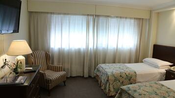 A hotel room with two beds, a desk with a lamp, a chair, and a TV mounted on the wall.