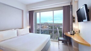 Habitación de hotel con una cama grande, un escritorio con una silla, un televisor y vistas a la ciudad a través de amplias ventanas.