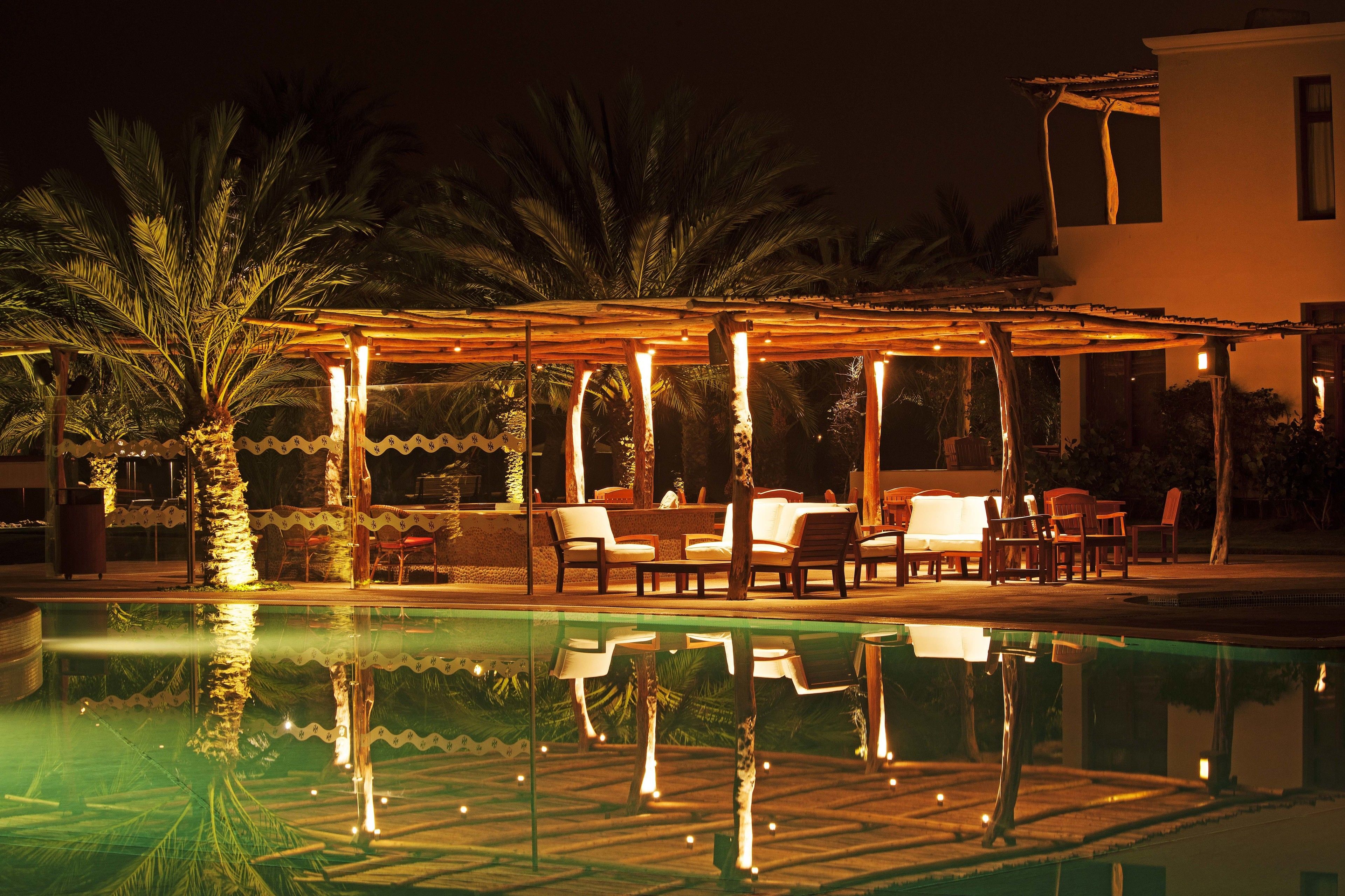 A nighttime pool area with a covered seating area, palm trees, and a reflection in the water.
