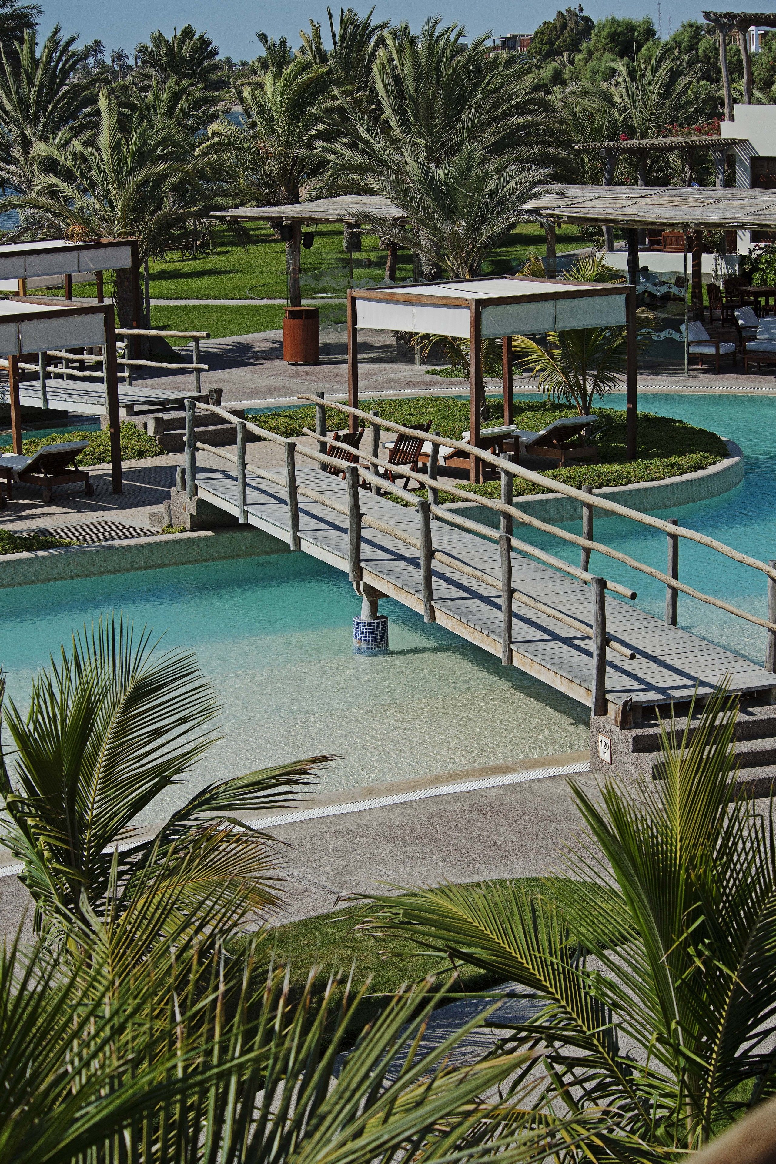 A pool area with a wooden bridge, lounge chairs, and palm trees.