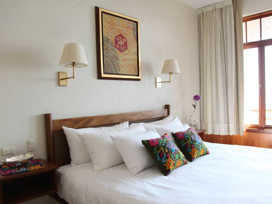 Una habitación de hotel con una cama grande, dos lámparas de noche, una mesita de noche, una obra de arte enmarcada, una ventana con cortinas y un jarrón con una flor.