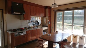 A kitchen with wooden cabinets, a gas stove, a sink, and a window with a view of greenery. - Lilac Hotels and Resort (Kanoya)