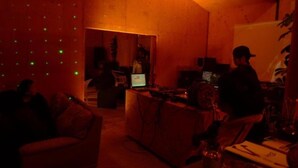 A dimly lit room with wooden walls and a sloped ceiling. There is a laptop on a table, a chair, and a person standing in the background. - Lilac Hotels and Resort (Kanoya)