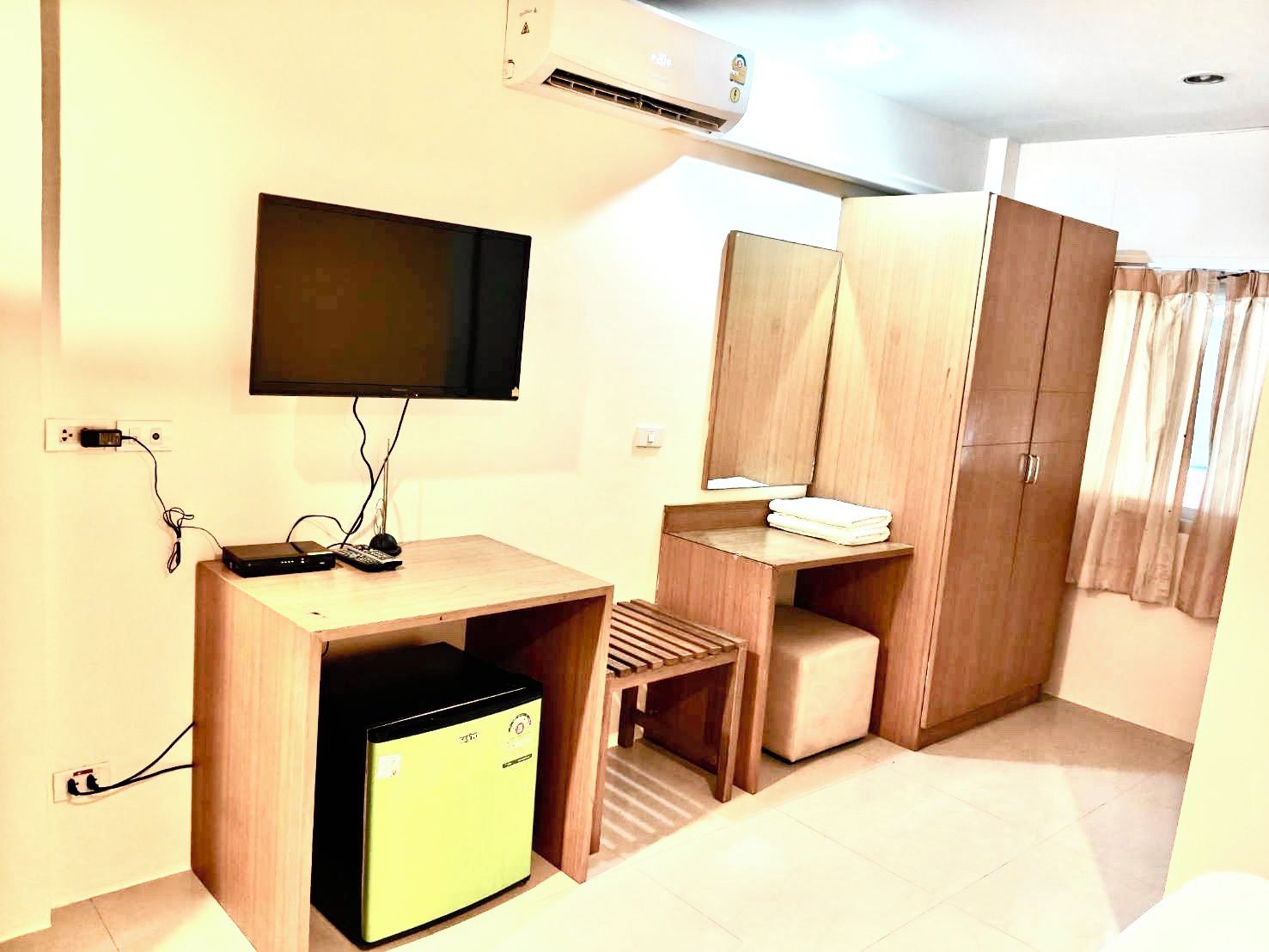 A compact room with a flat-screen TV mounted on the wall, a small wooden desk with a computer and keyboard, a mini-fridge, a mirror, and a wardrobe.