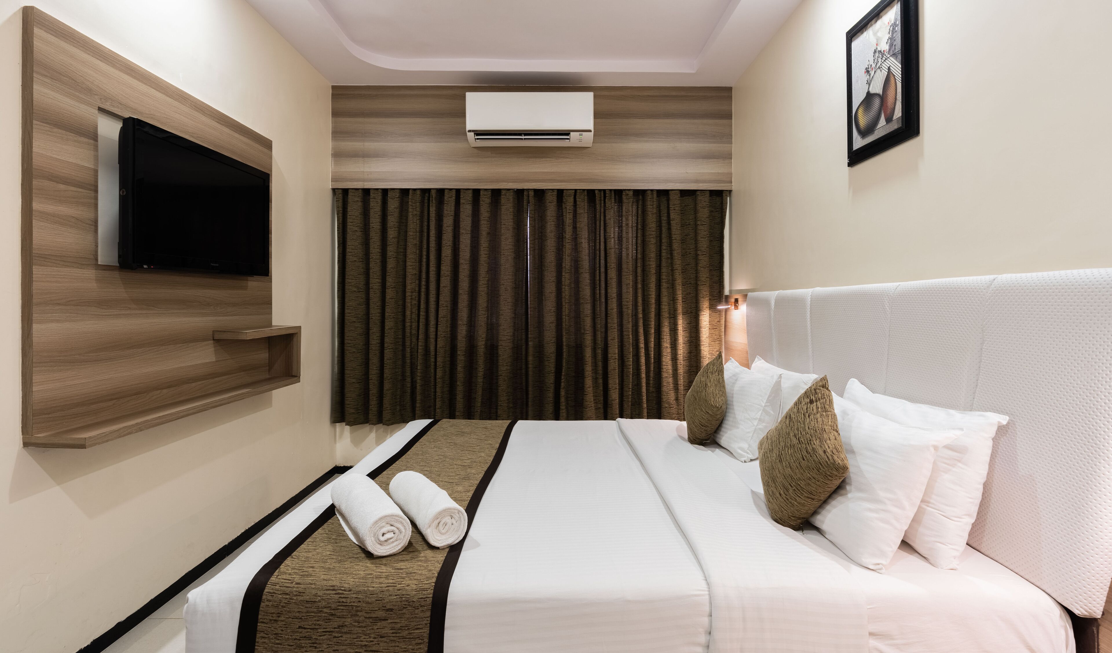 A hotel room with a flat-screen TV mounted on a wooden panel, a large air conditioning unit above the curtains, and a bed with white linens and textured pillows.