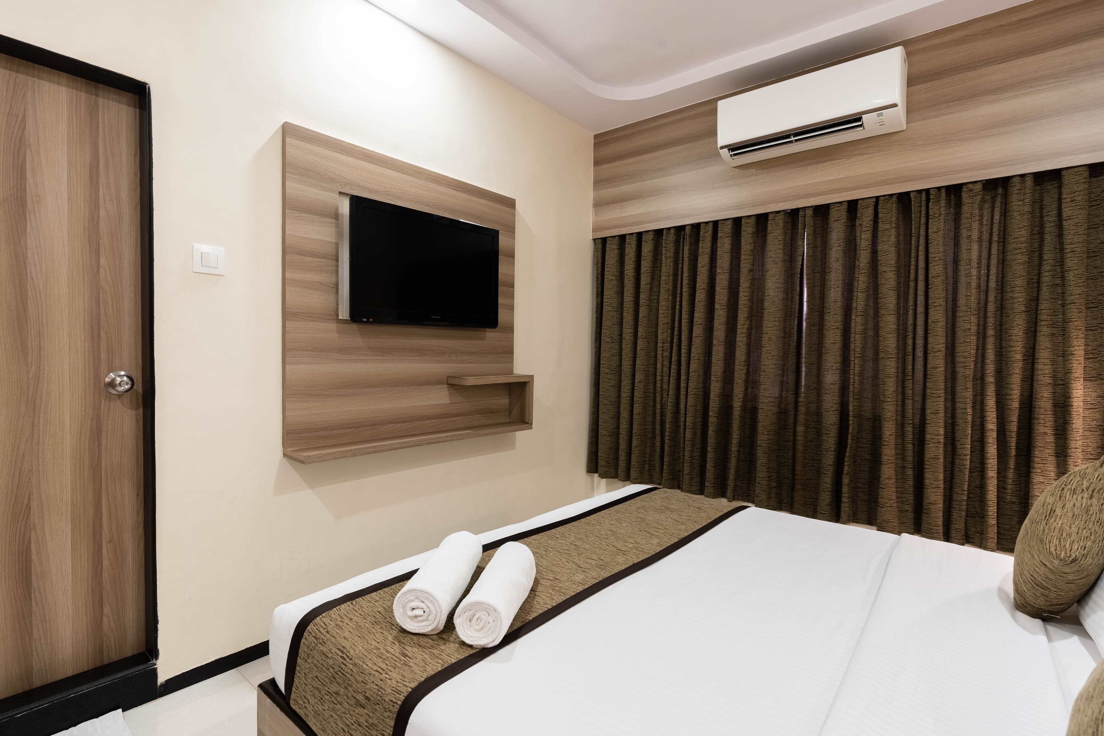 A hotel room with a bed, a mounted TV, an air conditioning unit, and a wooden door.