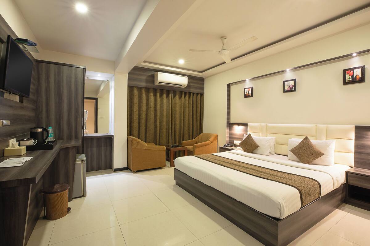 A hotel room with a bed, two armchairs, a desk with a television, and an air conditioning unit.