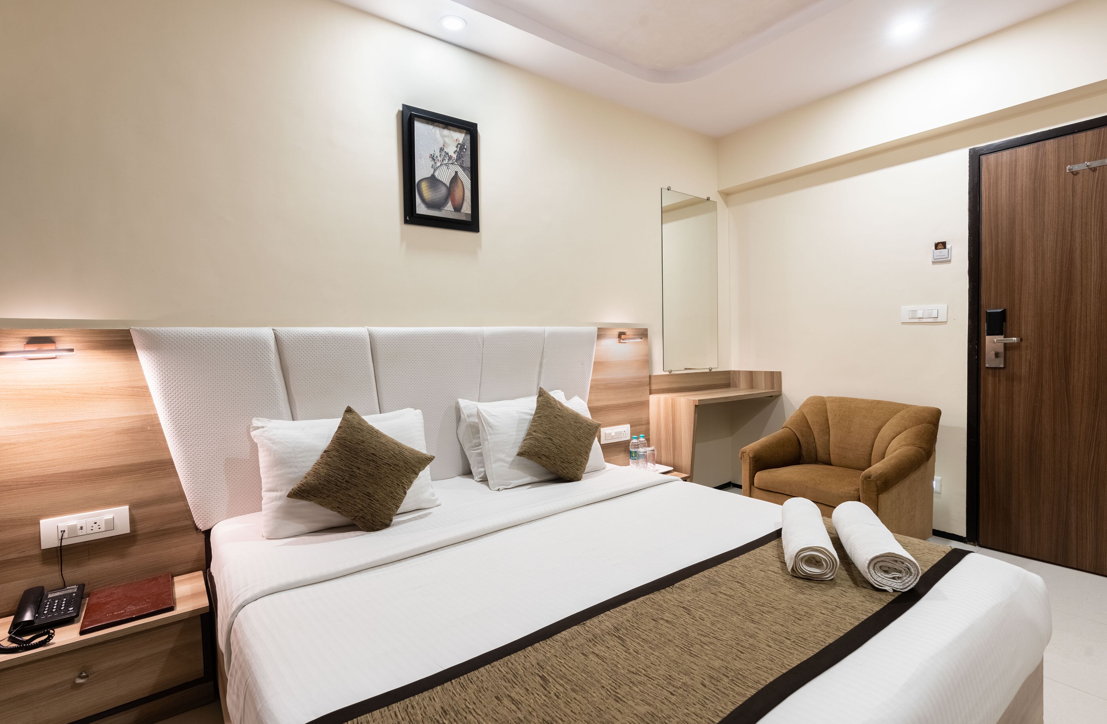 A hotel room with a large bed, a bedside table with a phone and a book, a chair, a mirror, and a wooden door.