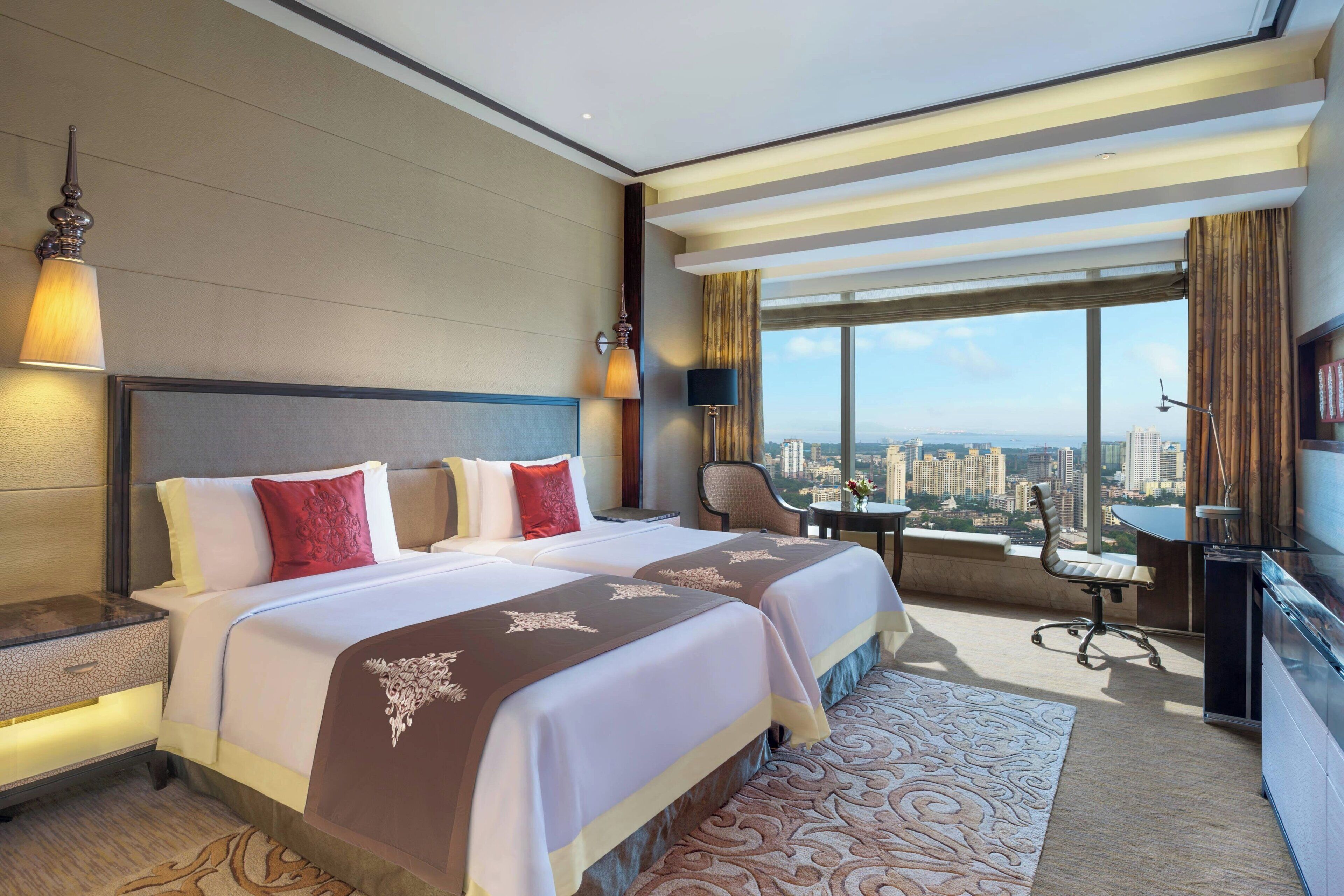 Habitación de hotel con dos camas, un escritorio, una silla y un amplio ventanal con vistas a la ciudad.