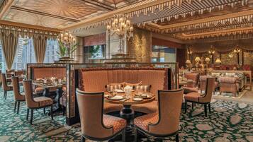 Um espaço de refeições luxuoso em hotel, com lustres ornamentados, estofados estampados e vista para a paisagem urbana.