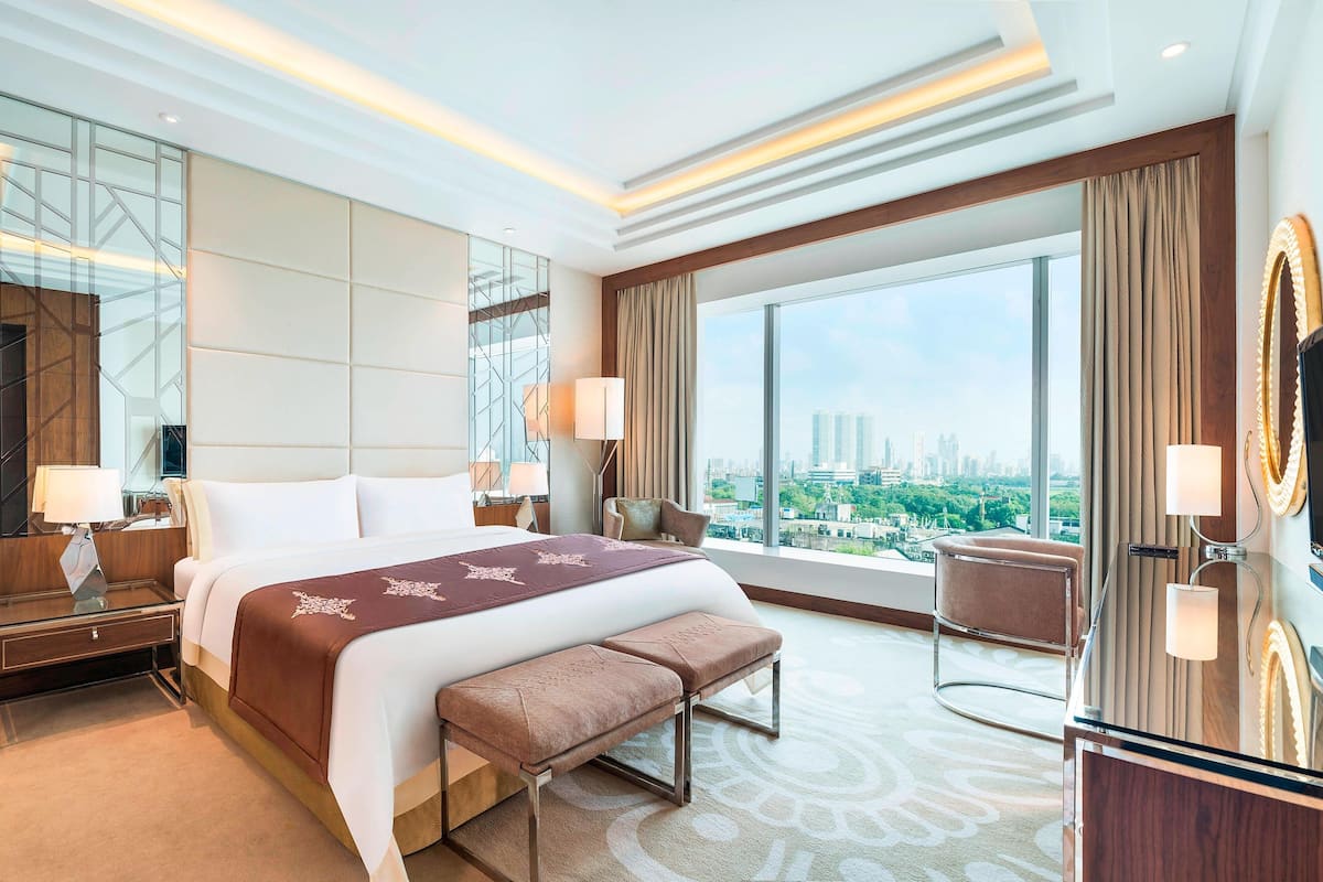 A modern hotel room with a large bed, a bench, a vanity with a mirror, and a view of the city.