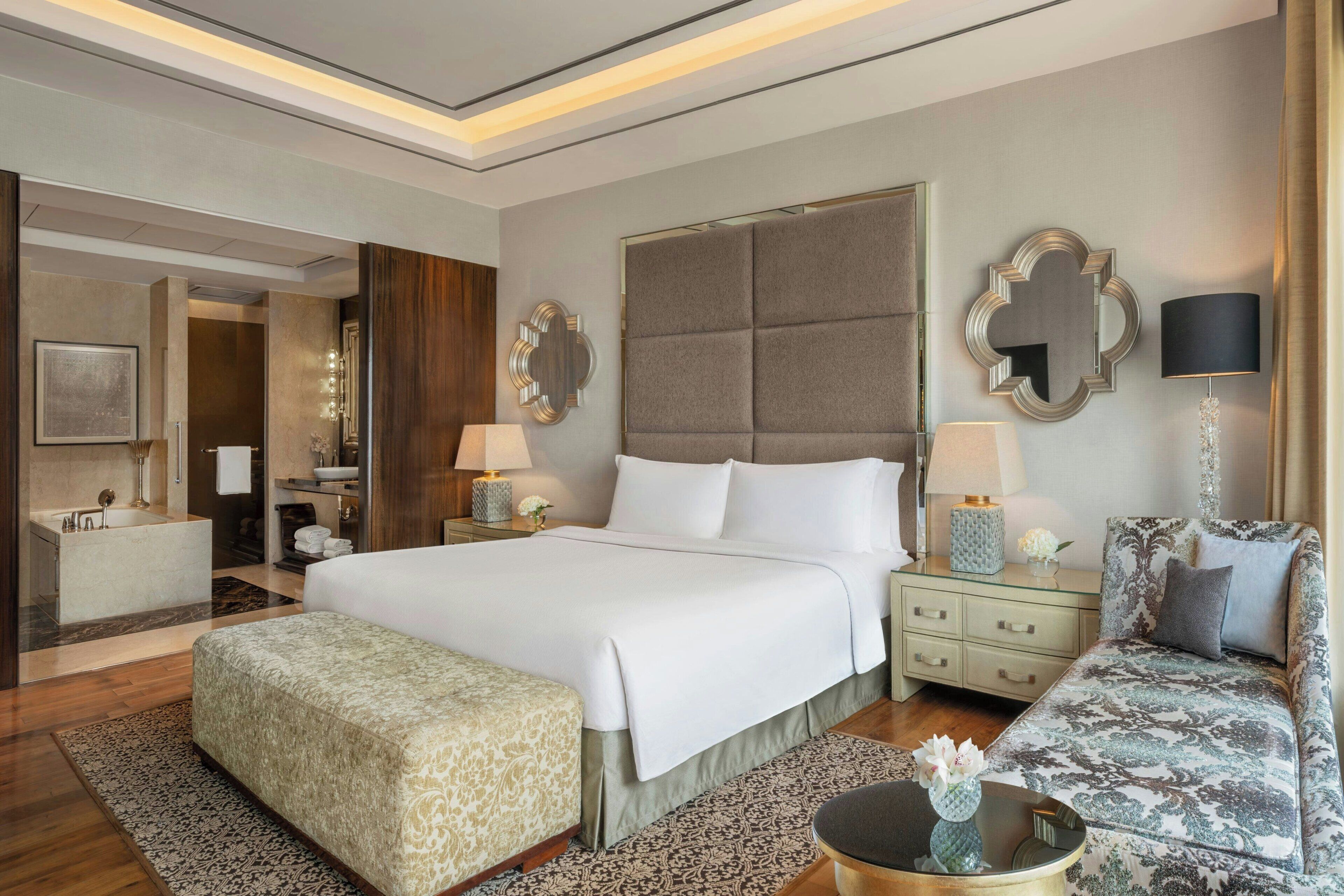 A hotel room with a large bed, a sofa, a nightstand, a mirror, and a bathroom visible through an open door.